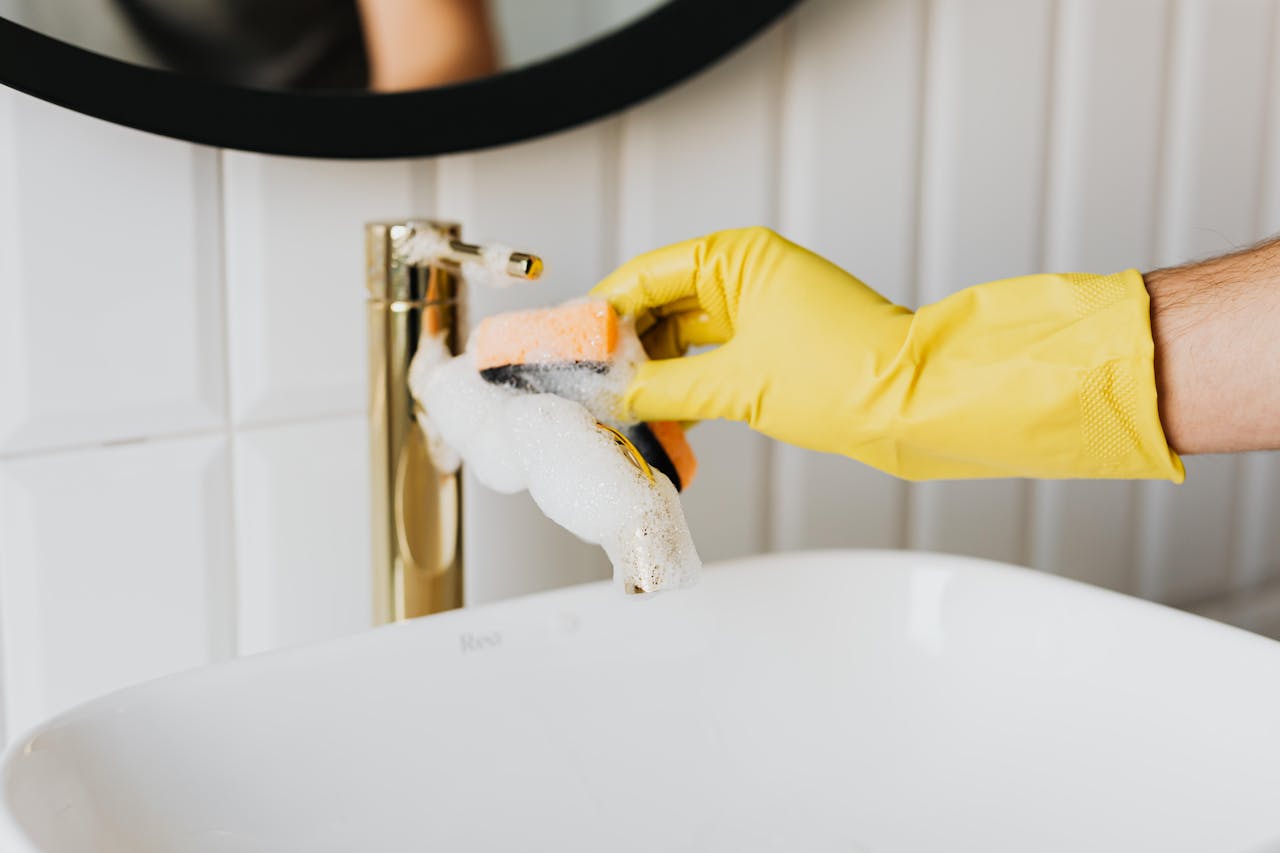 Closeup of a hand in a yellow glove cleaning a bathroom faucet with a sponge and soap.